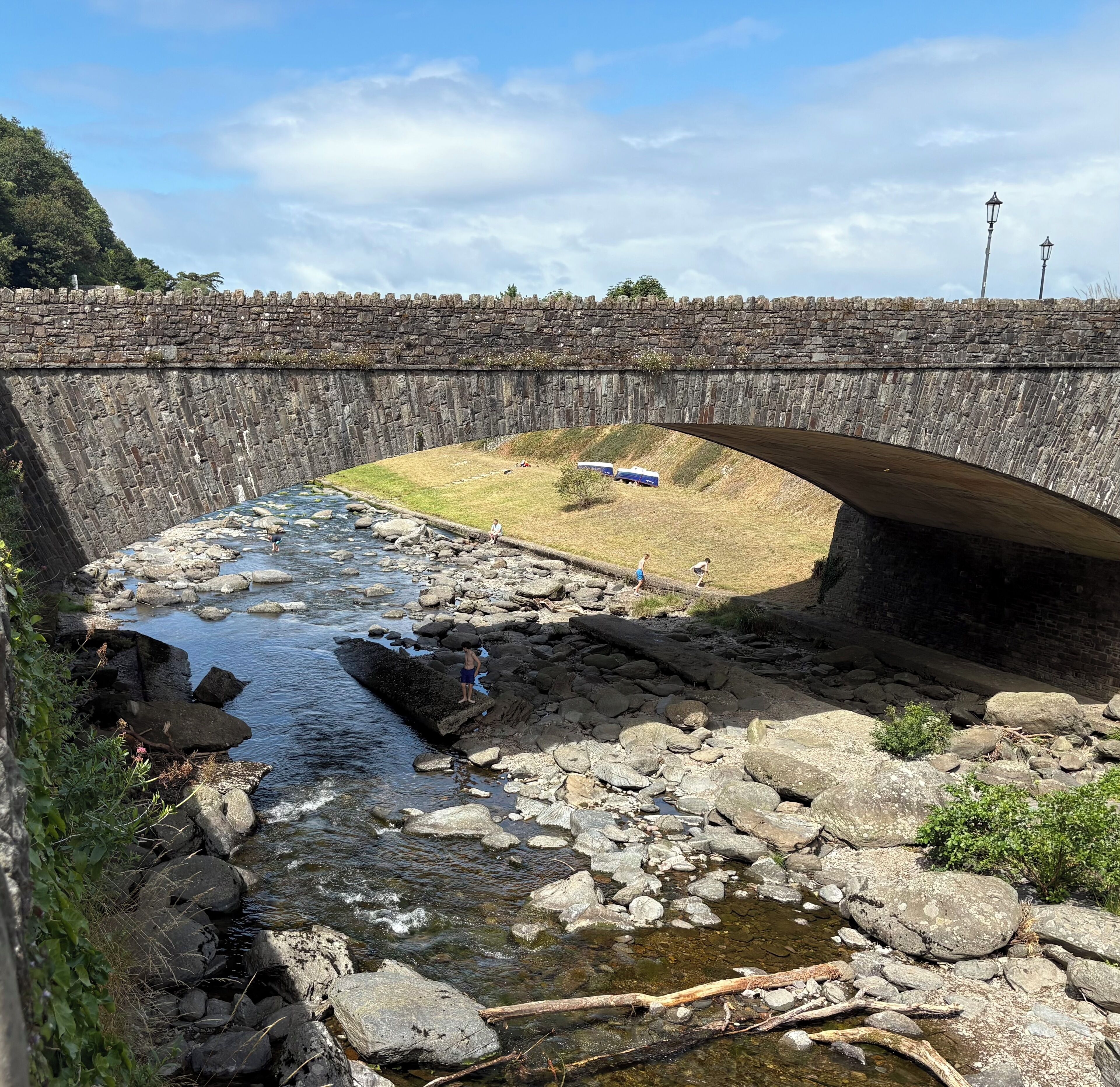 A stone bridge over a rocky stream with a grassy area and a few people.