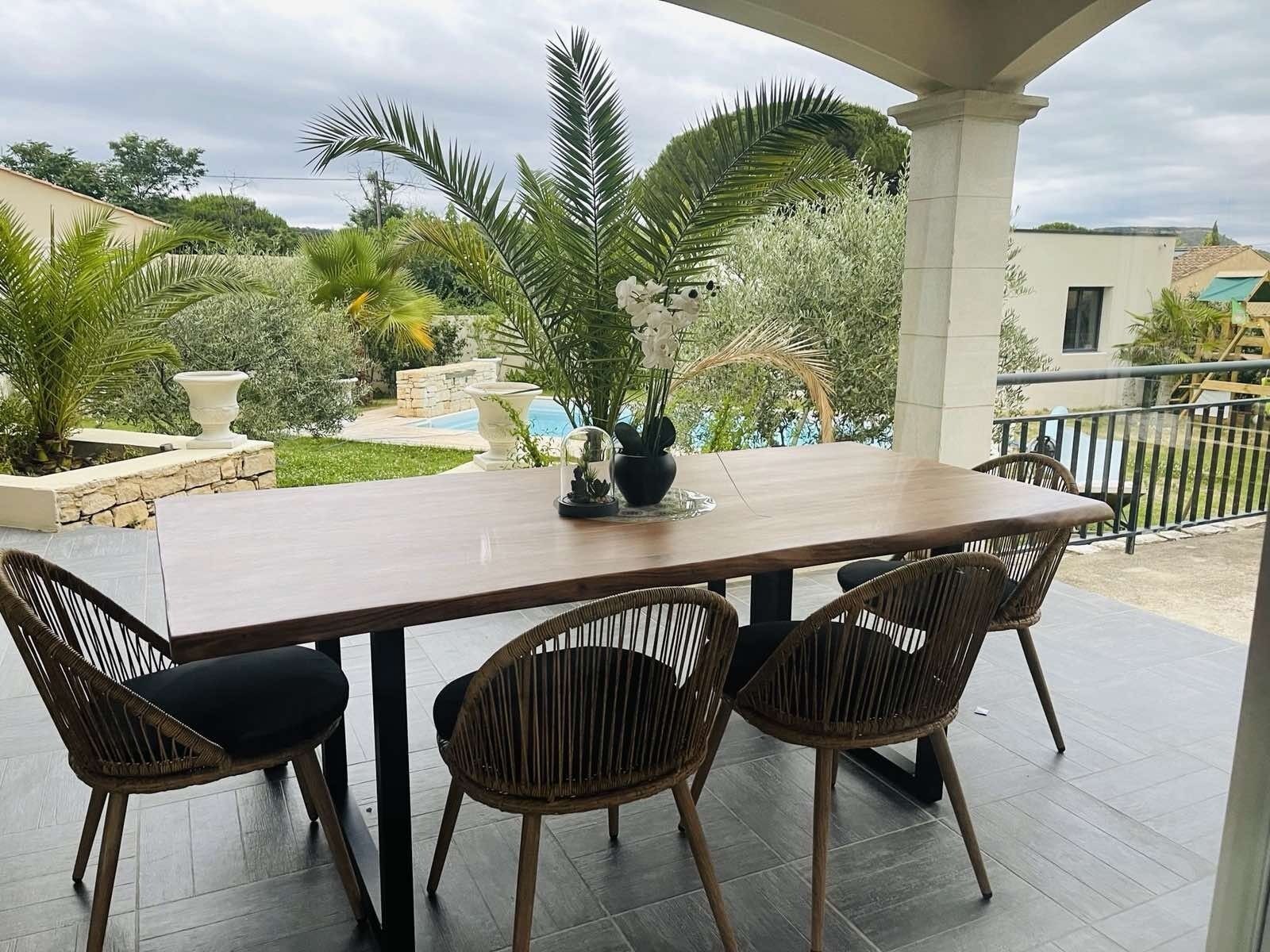 Um pátio externo com mesa de madeira e cadeiras de vime, com vista para um jardim ajardinado com palmeiras e uma piscina.