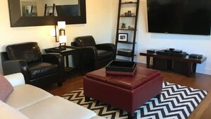 Ein modernes Wohnzimmer mit einem weißen Sofa, schwarzen Ledersesseln, einem roten Ottomanen, einem großen Spiegel, einem Fernseher und einem Holzbodenregal mit verschiedenen Gegenständen.