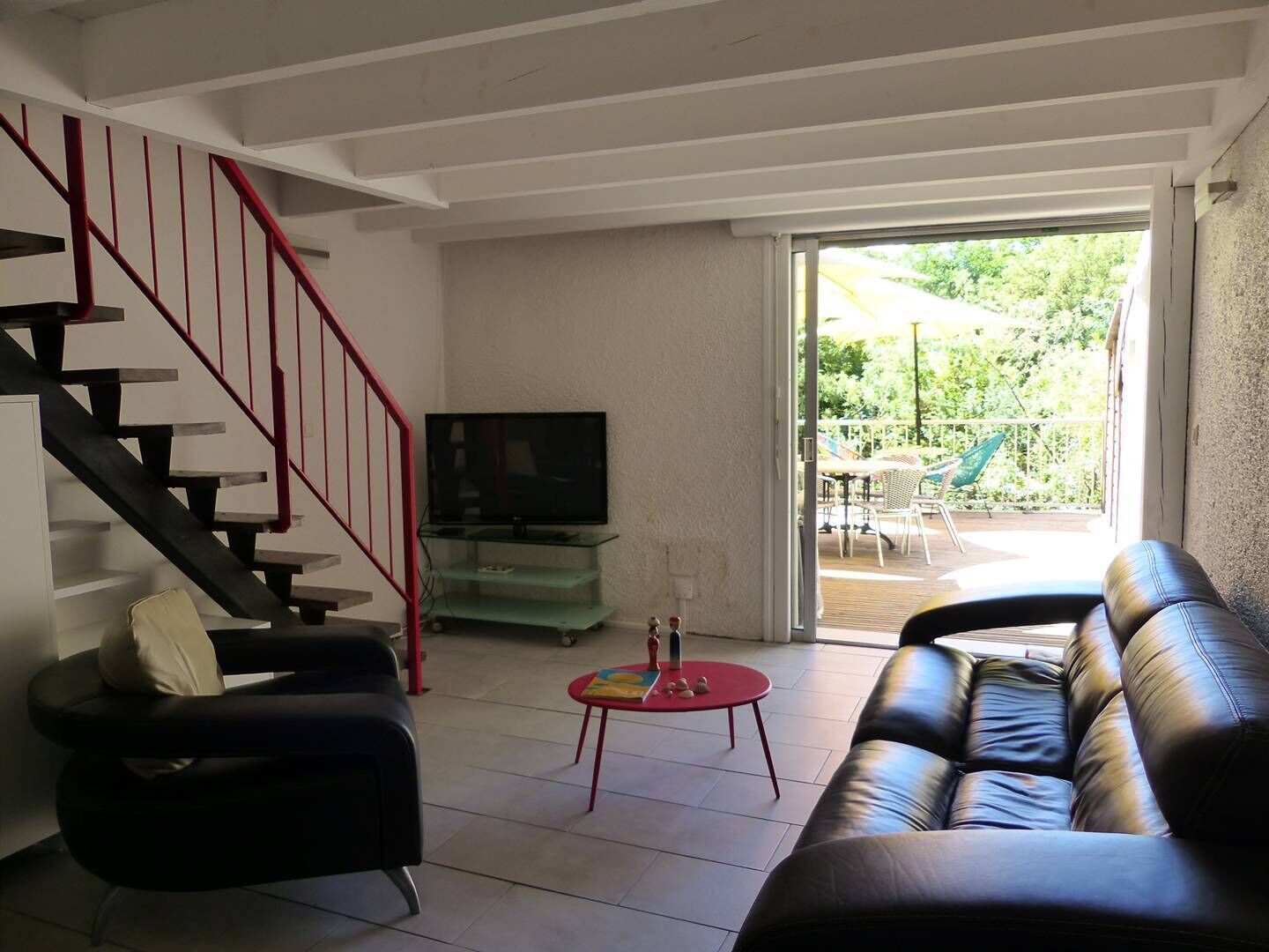 Un salon avec un escalier, un téléviseur posé sur un meuble en verre, un canapé, une petite table avec un pied rouge et une porte-fenêtre coulissante donnant sur un espace extérieur aménagé avec des chaises et des parasols.