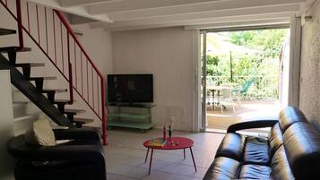 Un salon avec un escalier, un téléviseur posé sur un meuble en verre, un canapé, une petite table avec un pied rouge et une porte-fenêtre coulissante donnant sur un espace extérieur aménagé avec des chaises et des parasols.