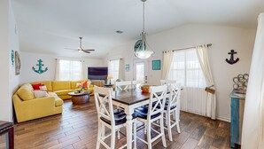 Una sala de estar con un sofá amarillo, una mesa de comedor con sillas blancas, un ventilador de techo y decoración temática náutica.