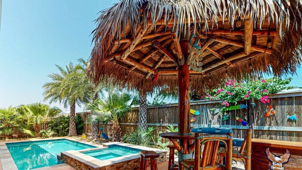 Zona de piscina con un jacuzzi, sillas de madera y una sombrilla de paja.