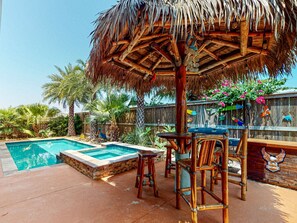 Zona de piscina con un jacuzzi, sillas de madera y una sombrilla de paja.
