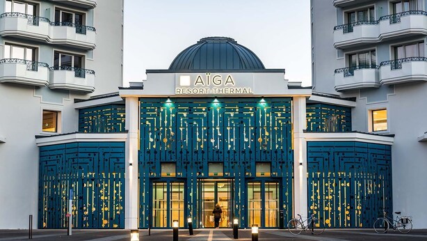 Ein Hotel mit Kuppel, einem Schild mit der Aufschrift 'AIGA RESORT THERMAL' und einer blauen Fassade mit vertikalen Linien.