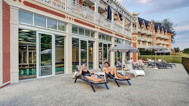 Un hotel con zona de piscina que incluye tumbonas y sombrillas, una entrada de vidrio amplia y un edificio de varios pisos con balcones.