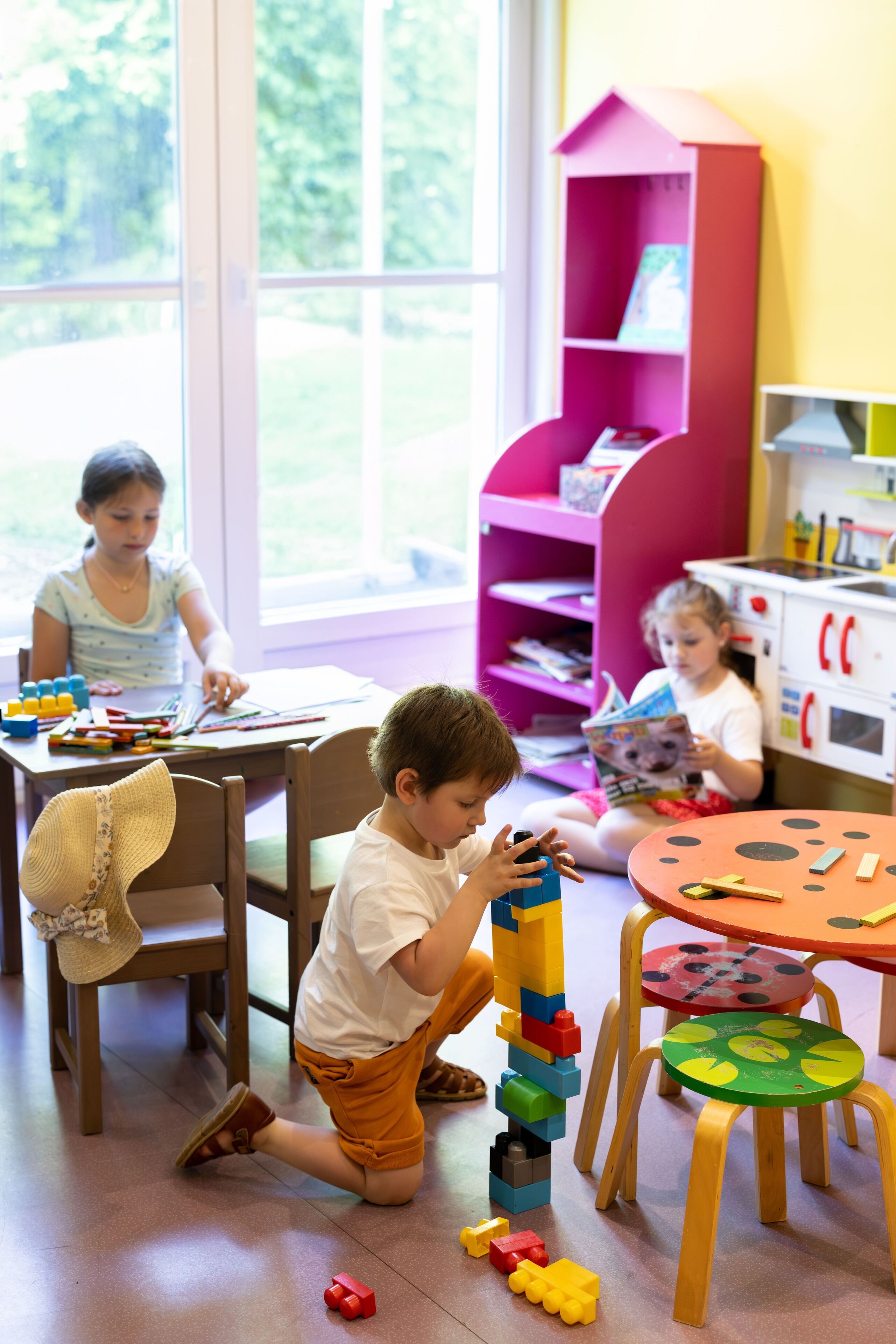 Een kind speelt met bouwblokken in een kamer met een groot raam, een roze boekenkast en een klein tafeltje met een tafelkleed met stippen.