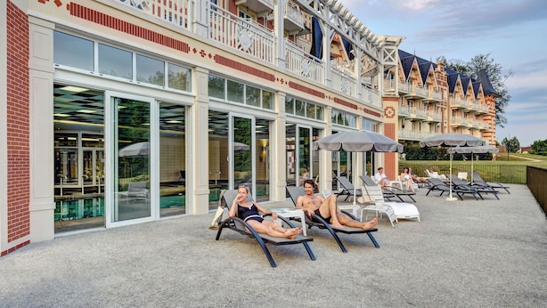 Un hotel con zona de piscina que incluye tumbonas y sombrillas, una entrada de vidrio amplia y un edificio de varios pisos con balcones.