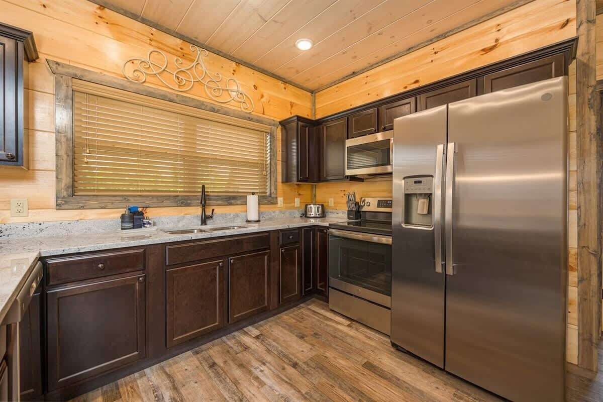 A modern kitchen with dark wooden cabinets, stainless steel appliances, and a large window with blinds.