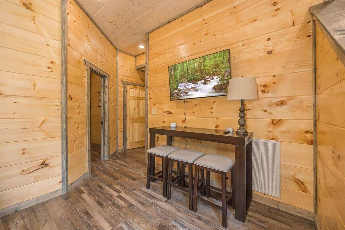Interior de madera con un pasillo, un banco con taburetes, una mesa con lámpara y un televisor en la pared.