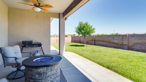 Un espacio de patio cubierto con un hogar a leña de piedra, un ventilador de techo y vista a un jardín con césped y una pared de ladrillo.
