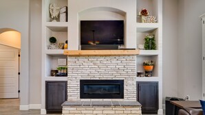 Un salón con chimenea de piedra, un televisor de pantalla plana montado encima y estantes con objetos decorativos.
