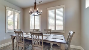 Un comedor con mesa y sillas de madera, ventanas amplias con persianas y una lámpara de araña.