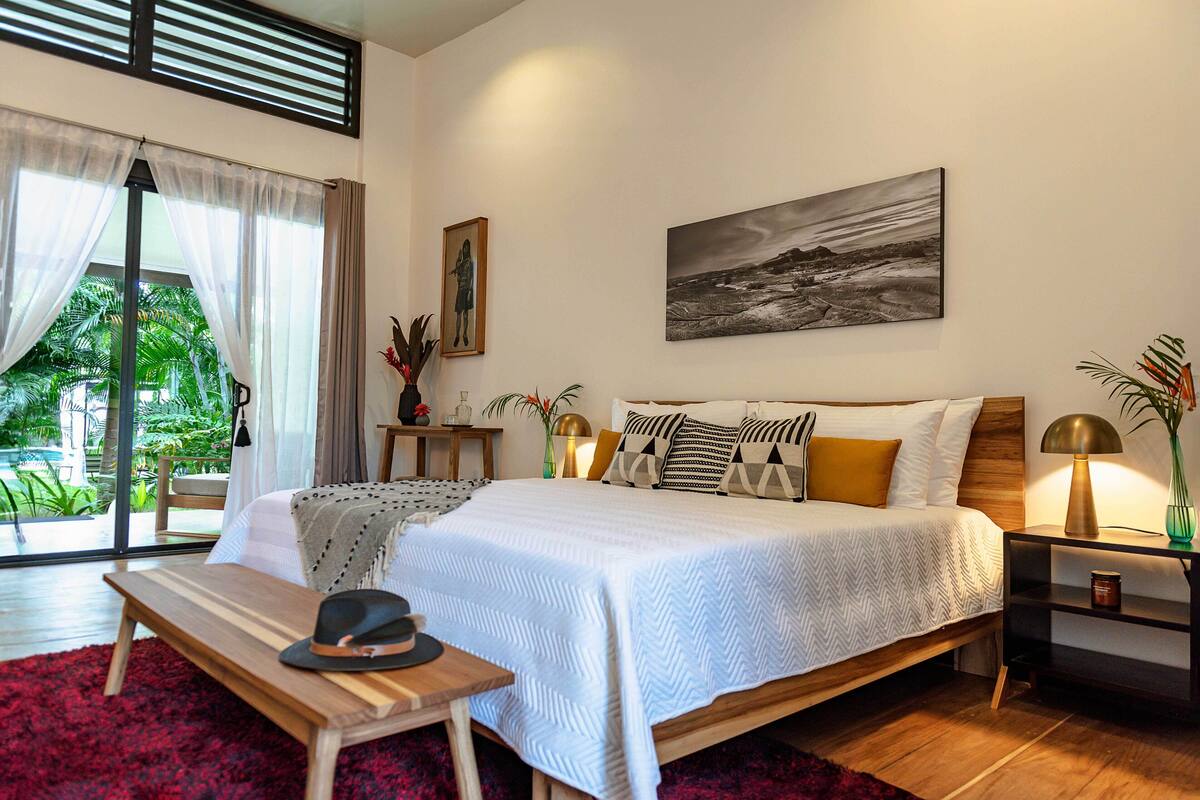 A modern bedroom with a large bed, a wooden bench, a nightstand, and a view of greenery outside.