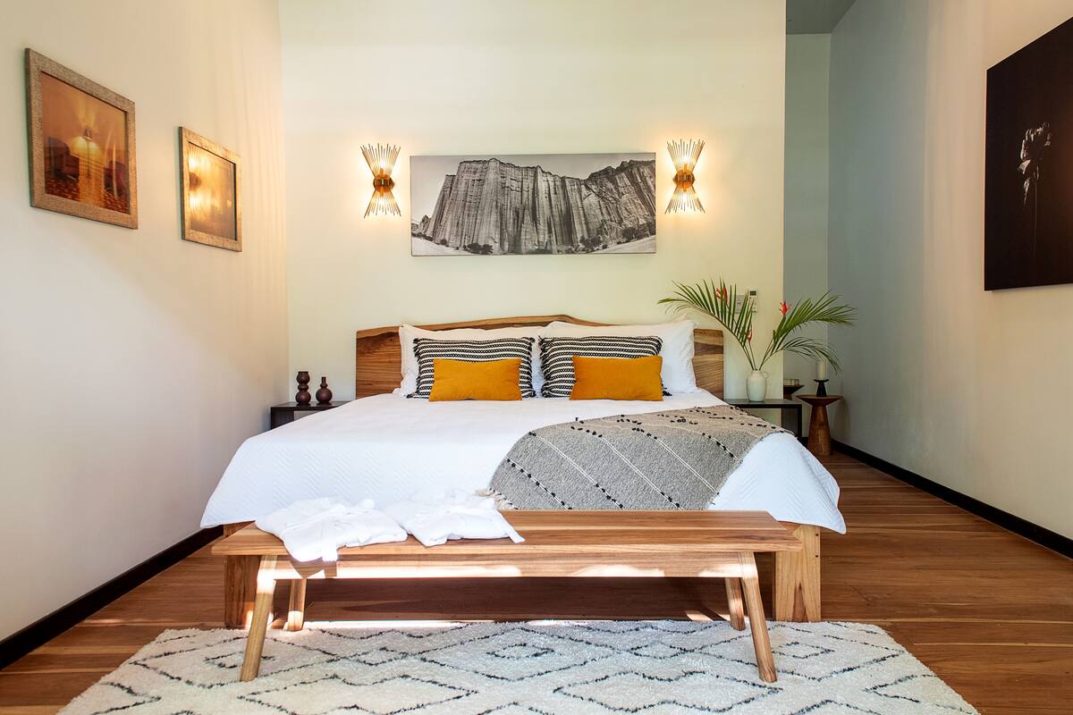 A bedroom with a large bed, wooden headboard, two wall-mounted lights, a framed picture, and a potted plant.