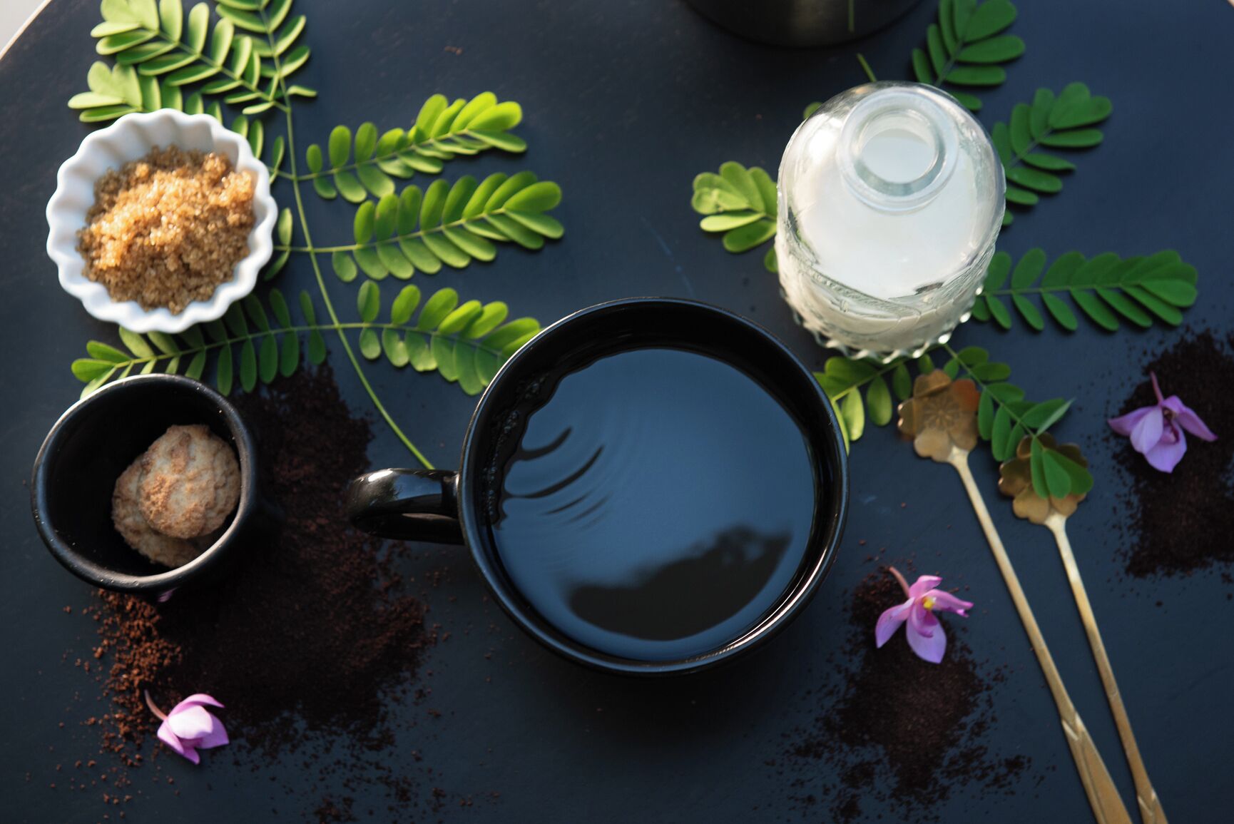 Une table sombre sur laquelle se trouvent une tasse noire, un bol blanc rempli de sucre brun, une bouteille en verre transparente, des feuilles vertes et une fleur rose.