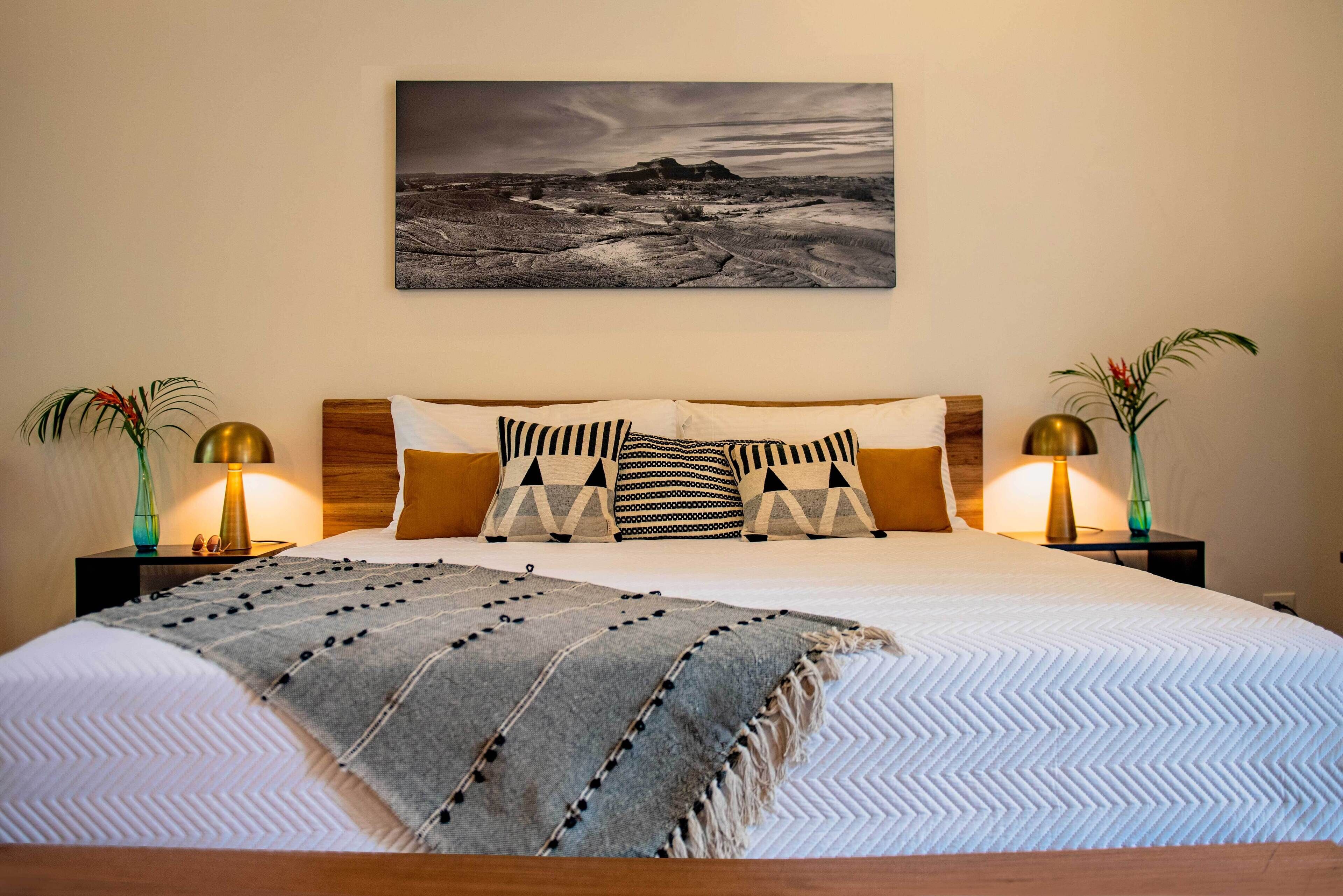 A neatly made bed with a patterned bedspread and pillows, flanked by two bedside tables with lamps and vases. A landscape painting is hung above the bed.
