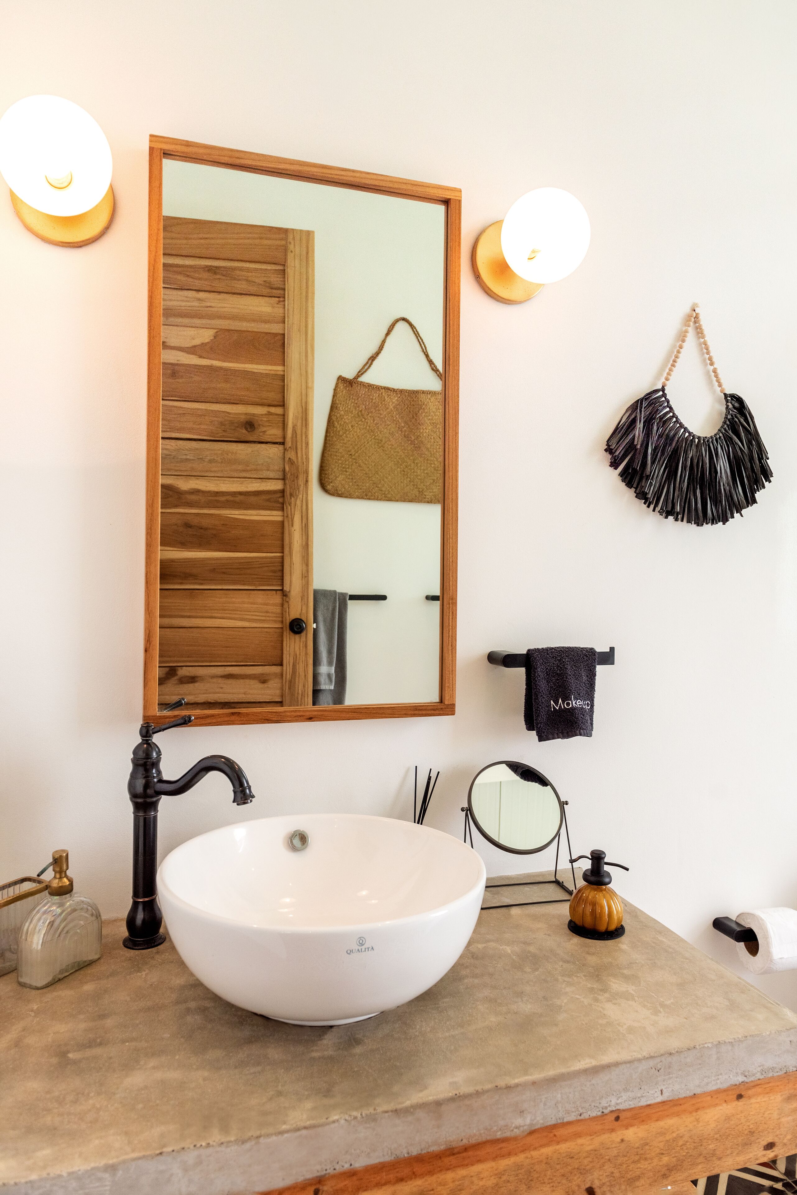 Une salle de bain avec une porte en bois, un lavabo blanc et un miroir. Il y a deux lampes fixées au mur et un porte-serviettes avec une serviette.