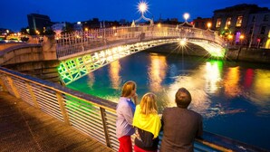 Un pont dont les garde-corps sont illuminés, surplombant une étendue d’eau, avec des personnes se tenant sur le trottoir adjacent.