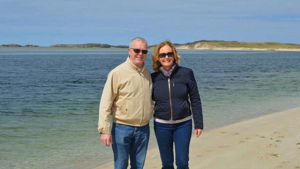 Un couple se tient sur une plage, avec vue sur la mer et sur des reliefs lointains.