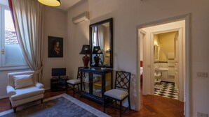 Een woonkamer met een witte fauteuil, een donkere console tafel met decoratieve objecten, een grote spiegel en uitzicht op een badkamer met een geruit vloertje.