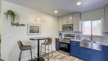 A modern kitchen with blue cabinets, a stainless steel sink, and a microwave. There is a round table with two chairs and a hanging light fixture.