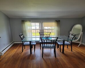Enjoy meals in this dining area with a glass-top table and four chairs, surrounded by a large window offering a view of the green lawn and a wooden fence.