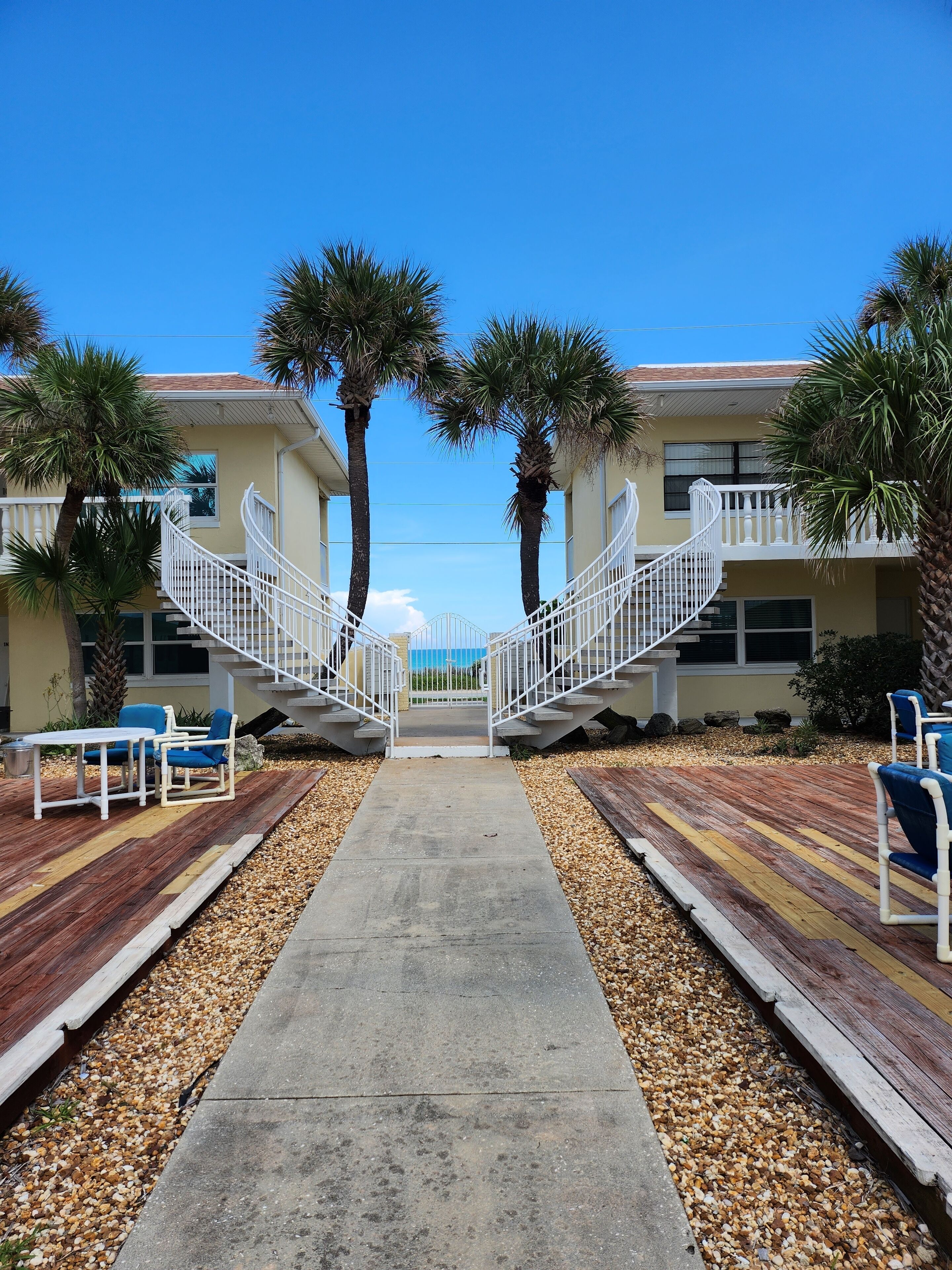 A hotel with a central concrete pathway leading to a set of white stairs, flanked by palm trees and outdoor seating areas.