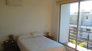 A bedroom with a bed, bedside tables, an air conditioner, and a large window with a view of buildings and greenery. - Zehn House in Girardot (Ricaurte)