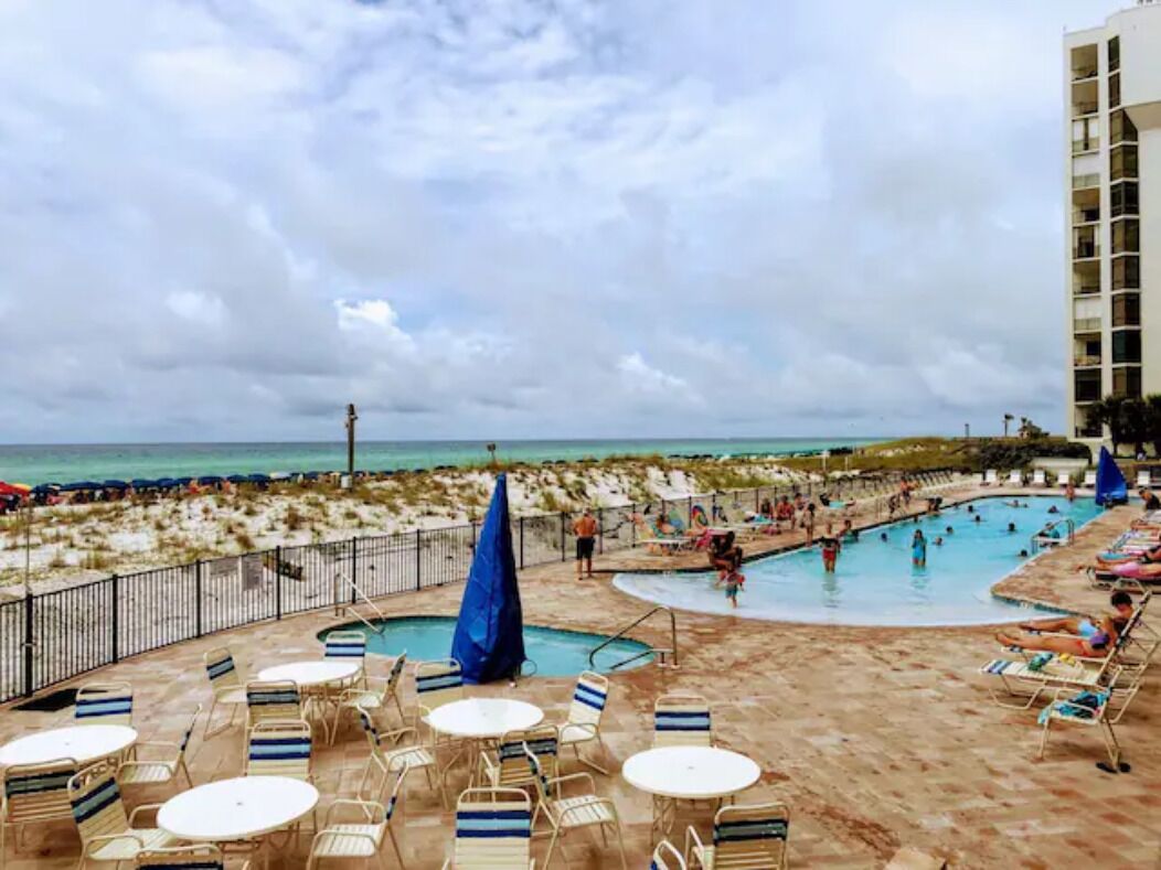 A pool area with lounge chairs and umbrellas, a beach in the background, and a building to the right.