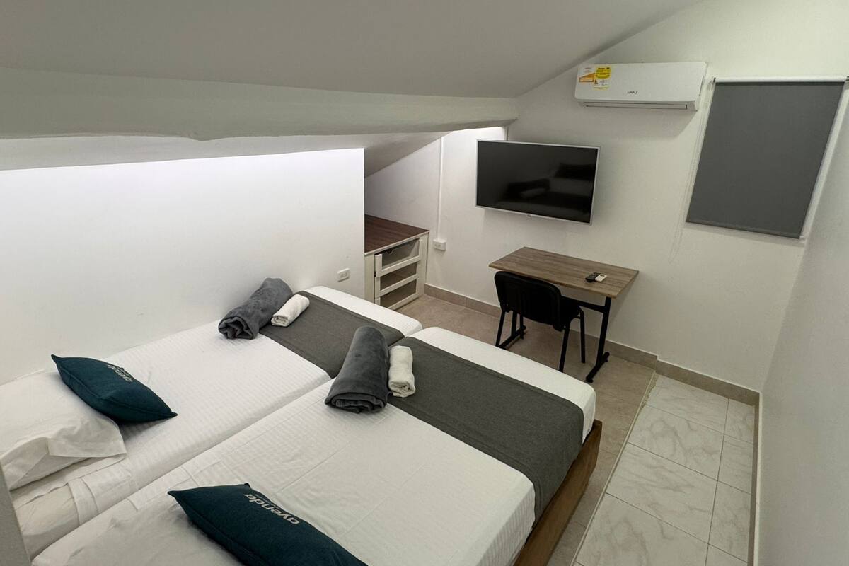 A compact hotel room with two beds, a desk, a TV, and a small kitchenette.