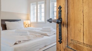 Una puerta de madera con manija de latón que da a un dormitorio con cama, ropa de cama blanca y una ventana con persianas.