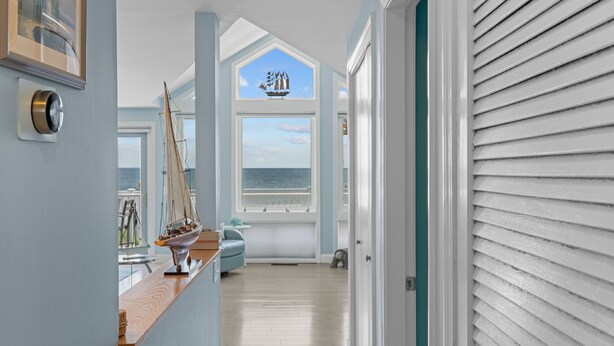 Ein Flur mit Blick auf den Ozean durch ein großes Fenster, ein Modell eines Segelbootes auf einem Holzregal und eine Tür mit weißen Fensterläden.