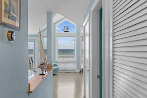 Un pasillo con vista al océano a través de un ventanal grande, un modelo de velero en un estante de madera y una puerta con contraventanas blancas.