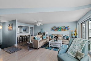 A spacious living room with a large window, a ceiling fan, and a kitchen area visible through an open door.