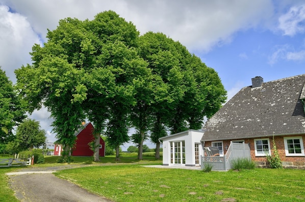Une maison de campagne avec un grand arbre devant, une grange rouge et un bâtiment blanc doté d’un toit en pente.