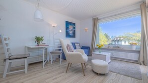 Un salón luminoso con suelo de madera, muebles blancos, un escritorio, una silla, un sofá y una ventana con cortinas.