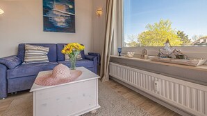 Una sala de estar moderna con un sofá azul, una mesita auxiliar blanca con un sombrero de sol, y una ventana con vista a árboles y un barco modelo.