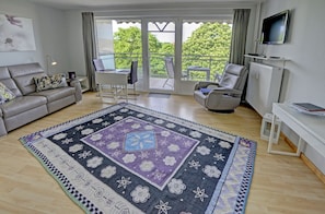 A living room with a sofa, a coffee table, a TV, a balcony with a chair, and a patterned rug.