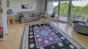 A modern living room with a grey sofa, a patterned rug, and a balcony with a view of greenery.