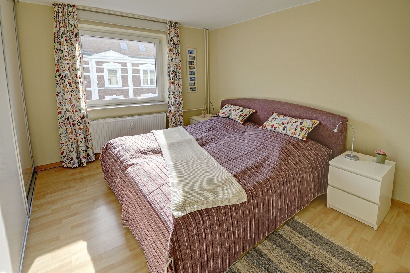 Un dormitorio con cama, mesita de noche, ventana con cortinas y radiador.