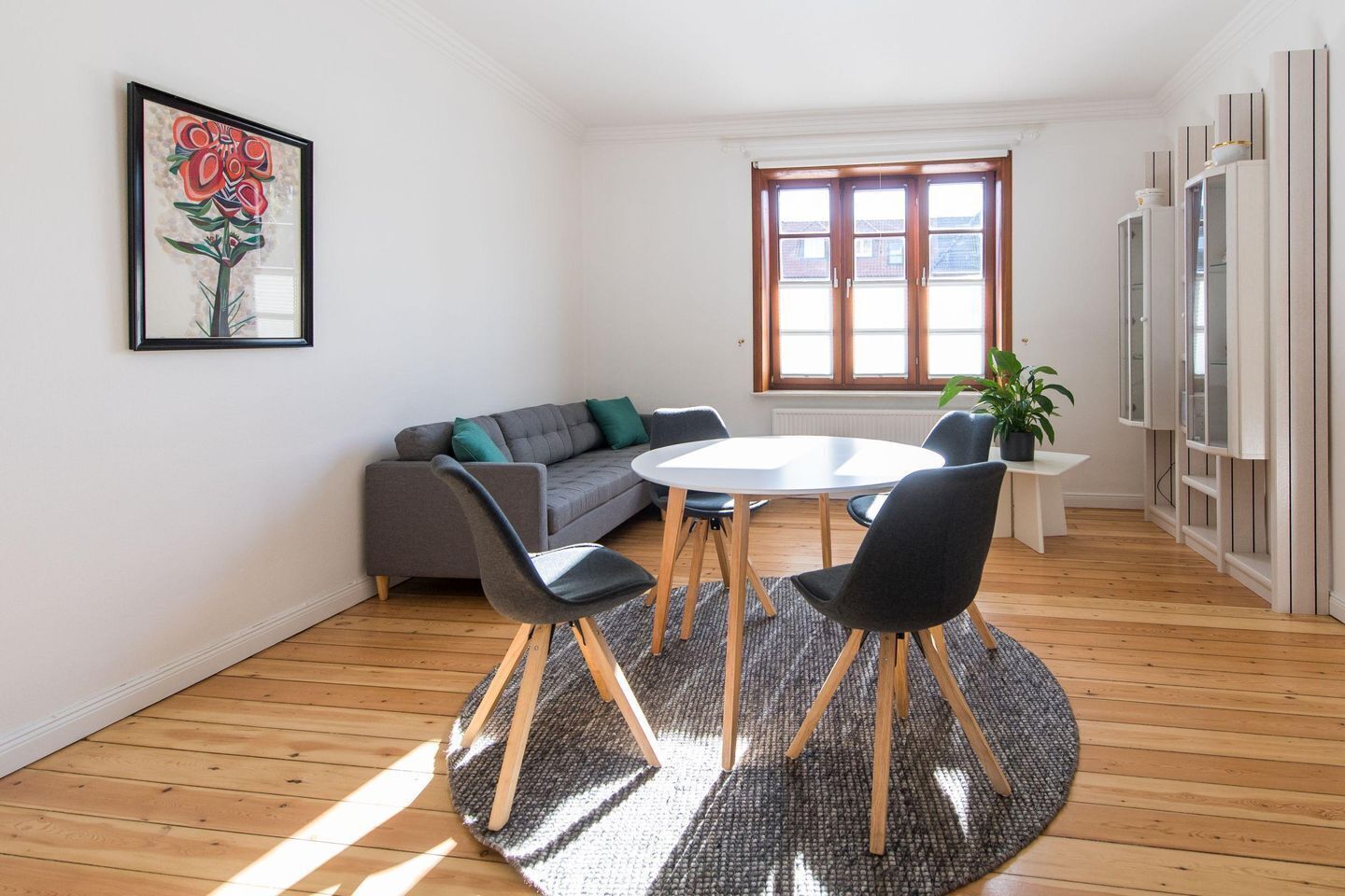 A modern living room with a grey sofa, a round table, and two chairs. There is a window with blinds, a potted plant, and a framed artwork on the wall.