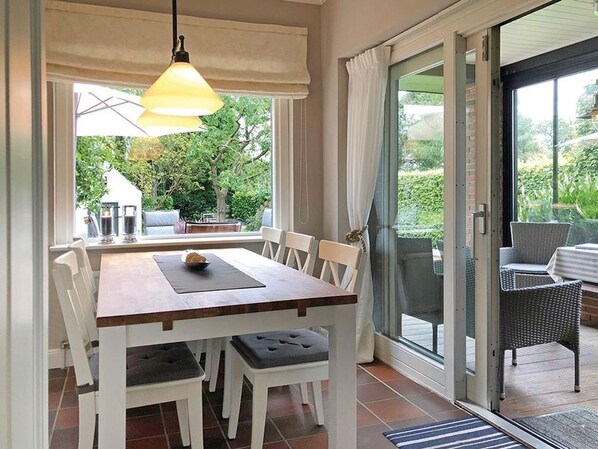 A dining area with a wooden table and white chairs, a hanging light fixture, and a view of the garden through a glass door.