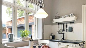 A kitchen with a dining table, chairs, a hanging light fixture, a window with a view, and a shelf with kitchen utensils. - fewo1846 - Home port Flensburg - fewo1846 - Home port Flensburg / elegant (Flensburg)