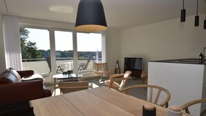 Una sala moderna con sofá, mesa de comedor y sillas, un balcón con vistas, y una zona de cocina con televisor y refrigerador.