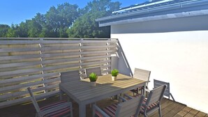 Una terraza de madera con una mesa, sillas, una planta en maceta y una pared blanca.