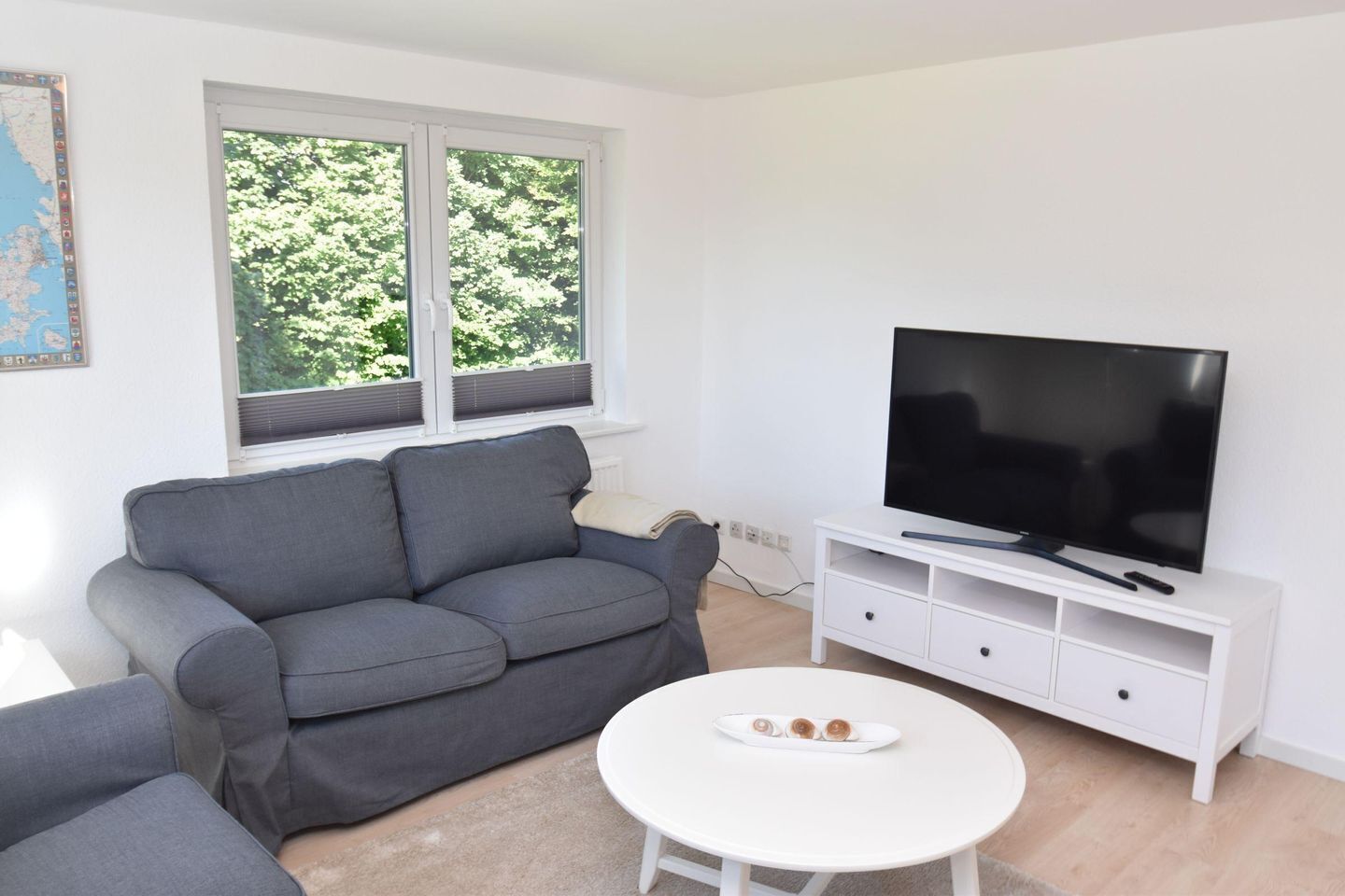 Uma sala de estar com um sofá cinzento, uma mesa de centro branca com um prato, uma TV sobre um suporte branco e uma janela com persianas.