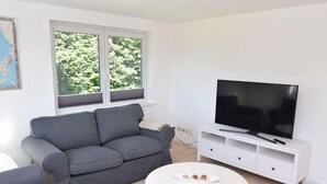 Una sala de estar con un sofá gris, una mesa de centro blanca con un plato, un televisor sobre un mueble blanco y una ventana con persianas.