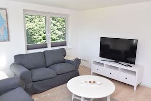 A living room with a grey sofa, a white coffee table with a plate, a TV on a white stand, and a window with blinds. - fewo1846 - Solskin - fewo1846 - Solskin / comfortable 3-room apartment in a rural location (Flensburg)