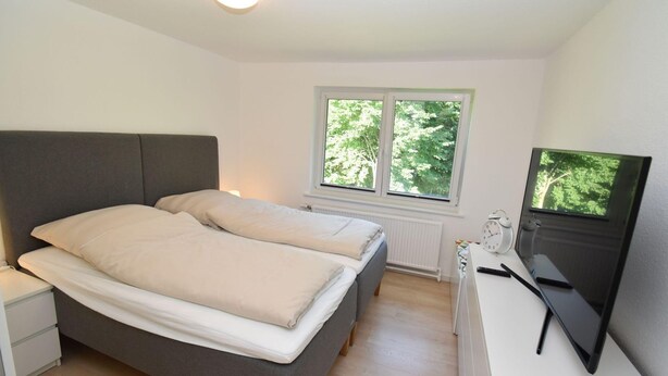 Un dormitorio con cama, mesitas de noche, un escritorio con computadora y ventana con vista a árboles.
